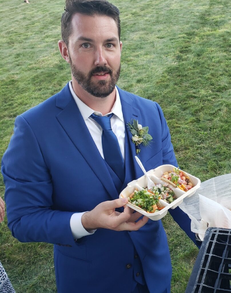 Man in blue suit and tie with a corsage on his lapel holding a tray of three tacos